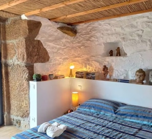 Une chambre avec un mur en pierre, un lit recouvert d’une couvre-lit à rayures bleues et blanches, une lampe de chevet et une étagère ornée d’objets décoratifs.