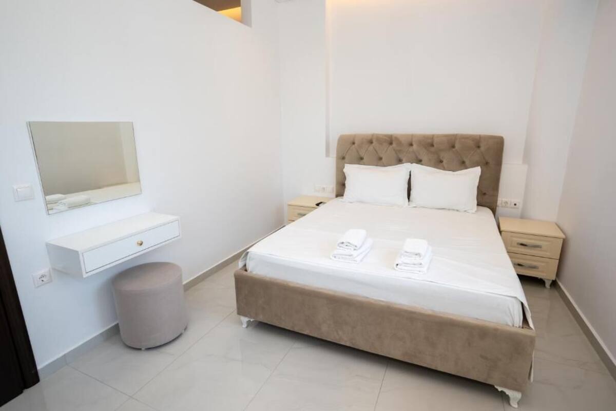 A bedroom with a bed, a bedside table, a nightstand, a mirror, a small stool, and a white wall unit.