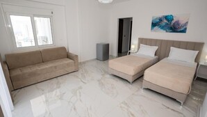 Une chambre d’hôtel avec un sol en marbre, un canapé beige, deux lits avec du linge de lit blanc, un haut-parleur gris et un tableau accroché au mur.