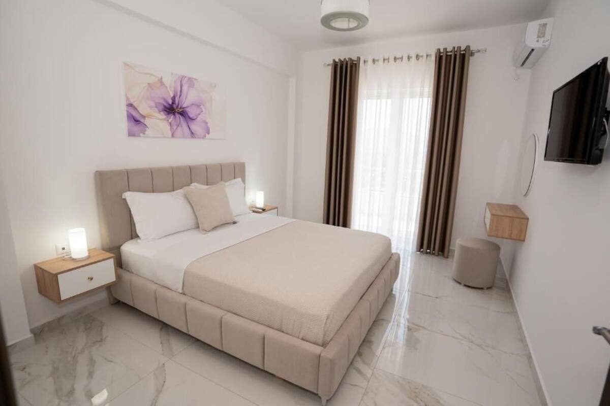 A bedroom with a bed, nightstands, a TV, a wall-mounted artwork, and a window with curtains.