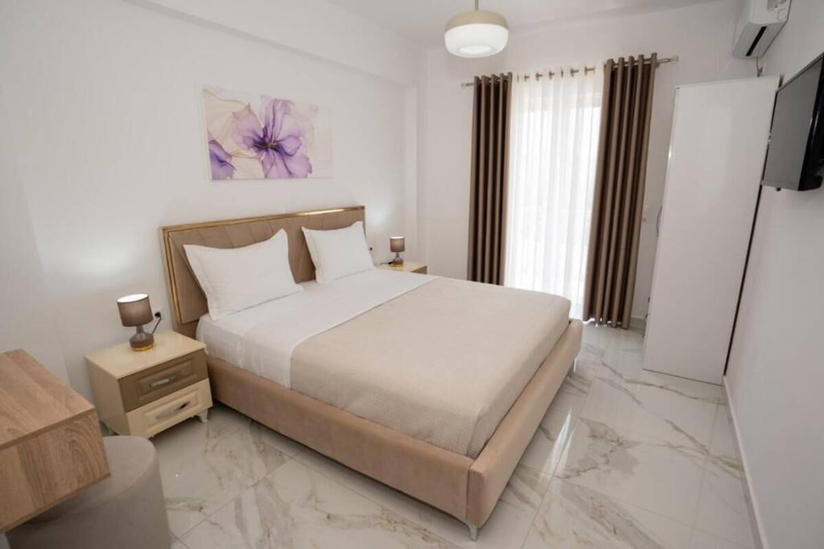 A hotel room with a bed, bedside tables, a TV, a window with curtains, and a wall with a floral painting.