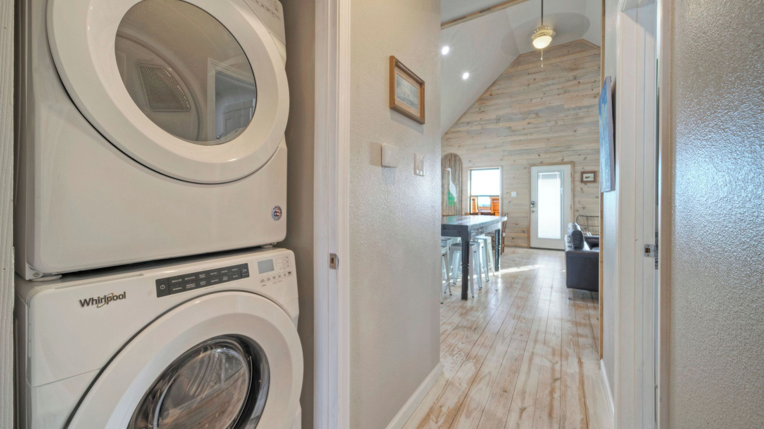 Une buanderie équipée de machines à laver et de sèche-linge Whirlpool superposées, avec un plancher en bois et une vue sur une salle à manger dotée d’un plafond haut et de poutres en bois.
