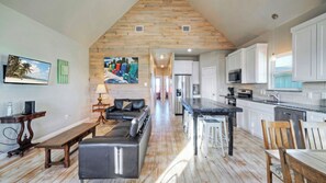 A spacious living area with a wooden ceiling, a black leather sofa, a wooden coffee table, and a kitchen with white cabinets and stainless steel appliances. - Pool, Boat Dock, Balcony w/ Bay Views Cooper's Hawk by Avantstay (Corpus Christi)