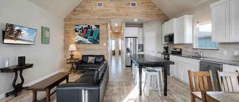 Un salón amplio con techo de madera, un sofá de cuero negro, una mesa de comedor y una cocina con armarios blancos y encimera de madera.