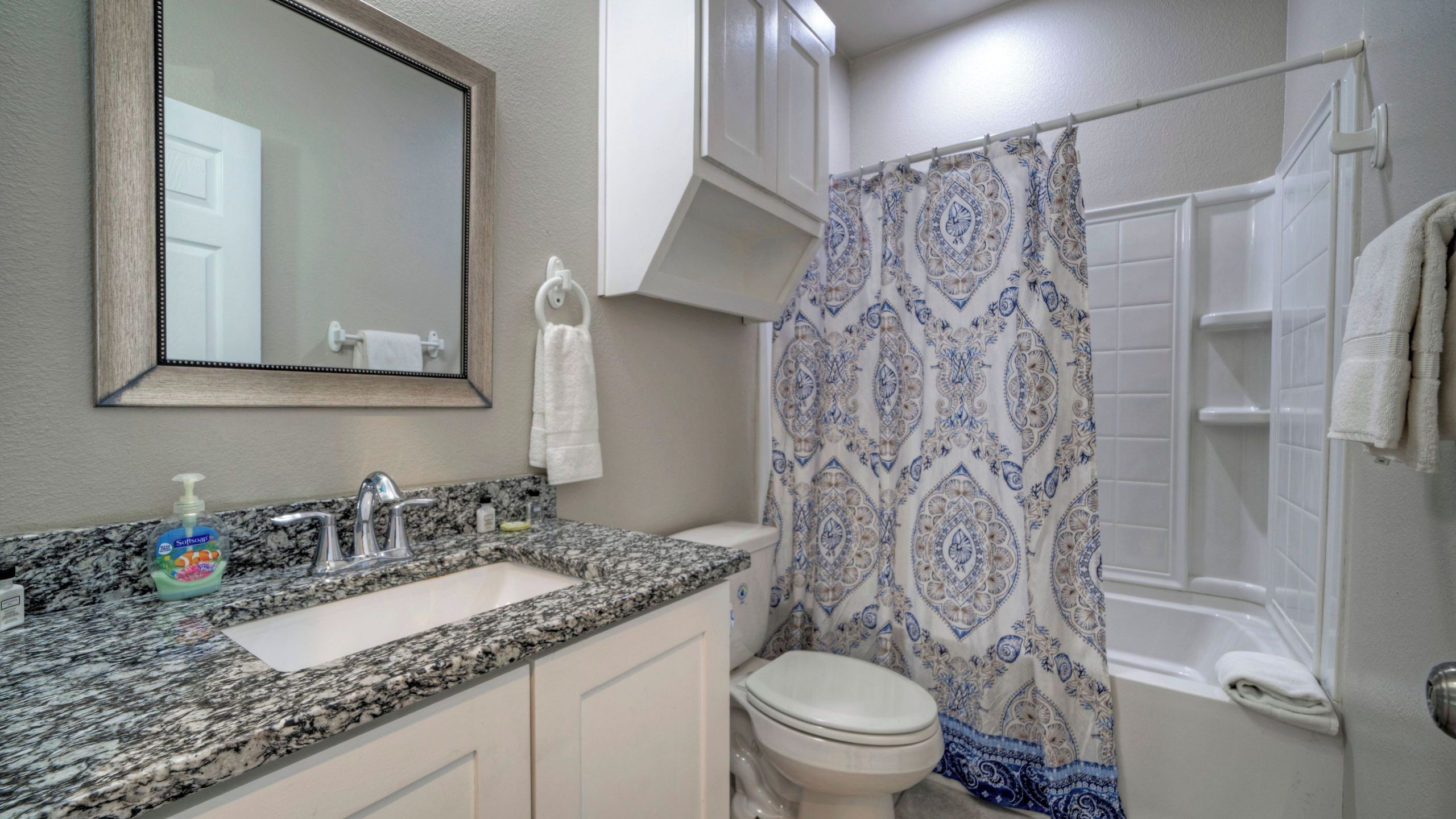 A bathroom with a granite countertop, a toilet, a shower with a patterned curtain, and a mirror above the sink.