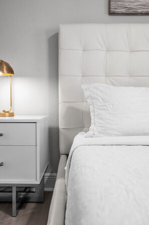 Un dormitorio con cama blanca, mesita de noche con lámpara y cabecero.