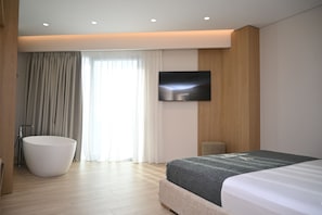 Una habitación de hotel moderna con una cama grande, televisión de pantalla plana, bañera y un amplio ventanal con cortinas.