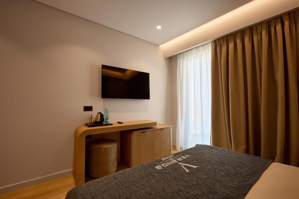 A modern hotel room with a bed, a flat-screen TV mounted on the wall, a wooden desk with a chair, and a window with curtains.