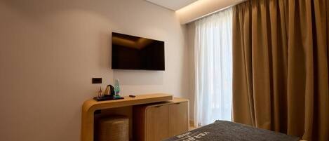A modern hotel room with a bed, a flat-screen TV mounted on the wall, a wooden desk with a chair, and a window with curtains.