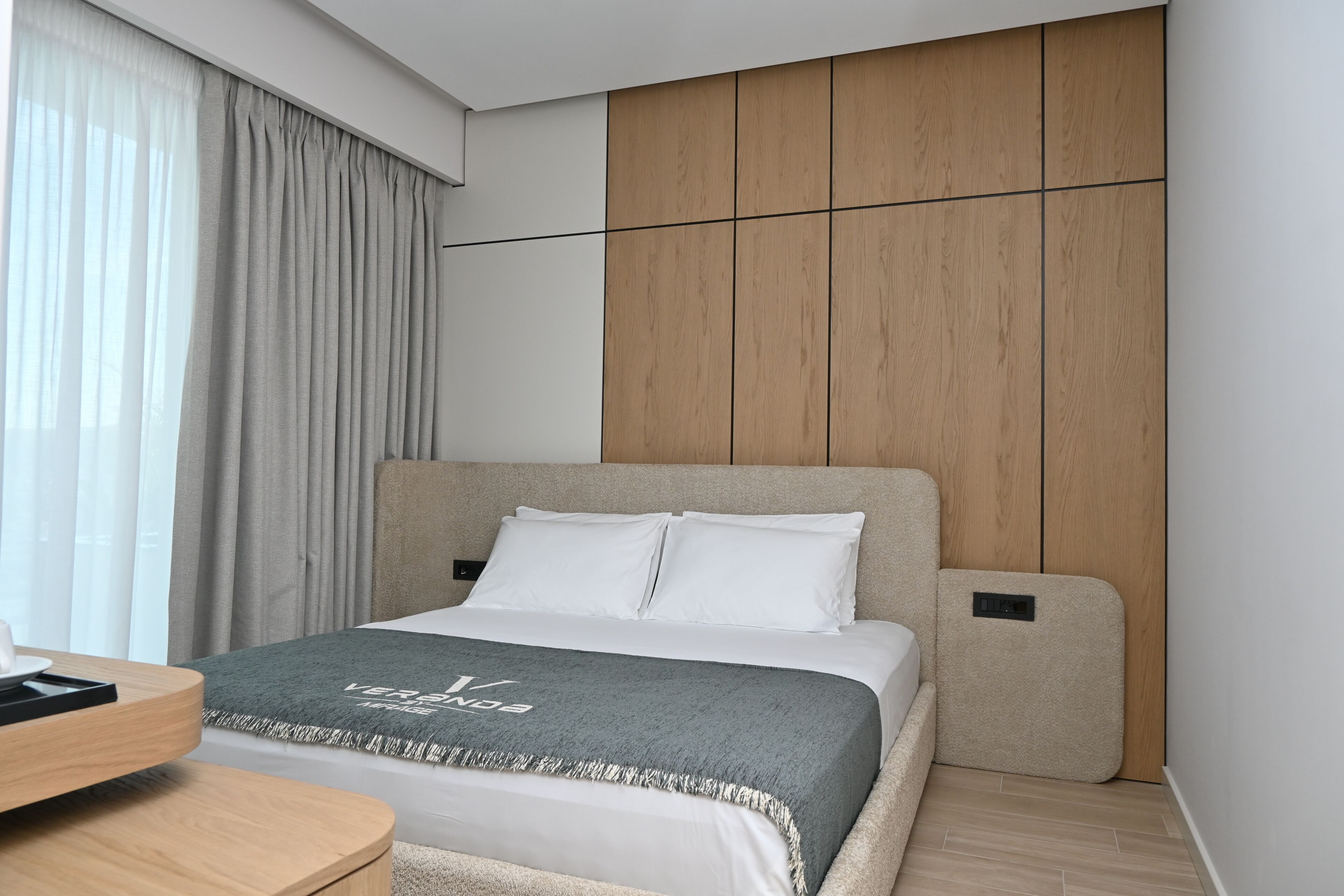 Una habitación de hotel moderna con cama, mesita de noche, escritorio y un amplio ventanal con cortinas.
