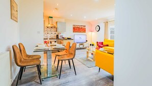 Un salon moderne avec une table à manger en verre, deux chaises, un canapé jaune, un téléviseur et une horloge au mur.