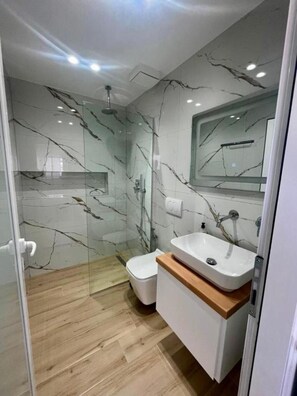 A modern bathroom with a glass shower enclosure, a white toilet, a wooden vanity with a sink, and marble-patterned walls. - Hotel Studio Eno Ksamil (Ksamil)