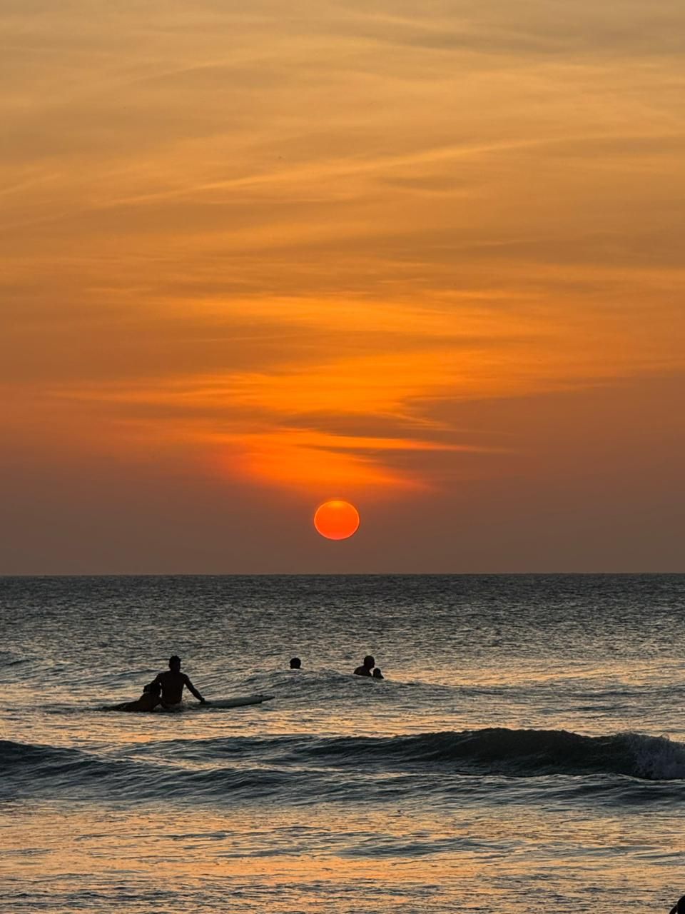 A sunset over the ocean with three people in the water.