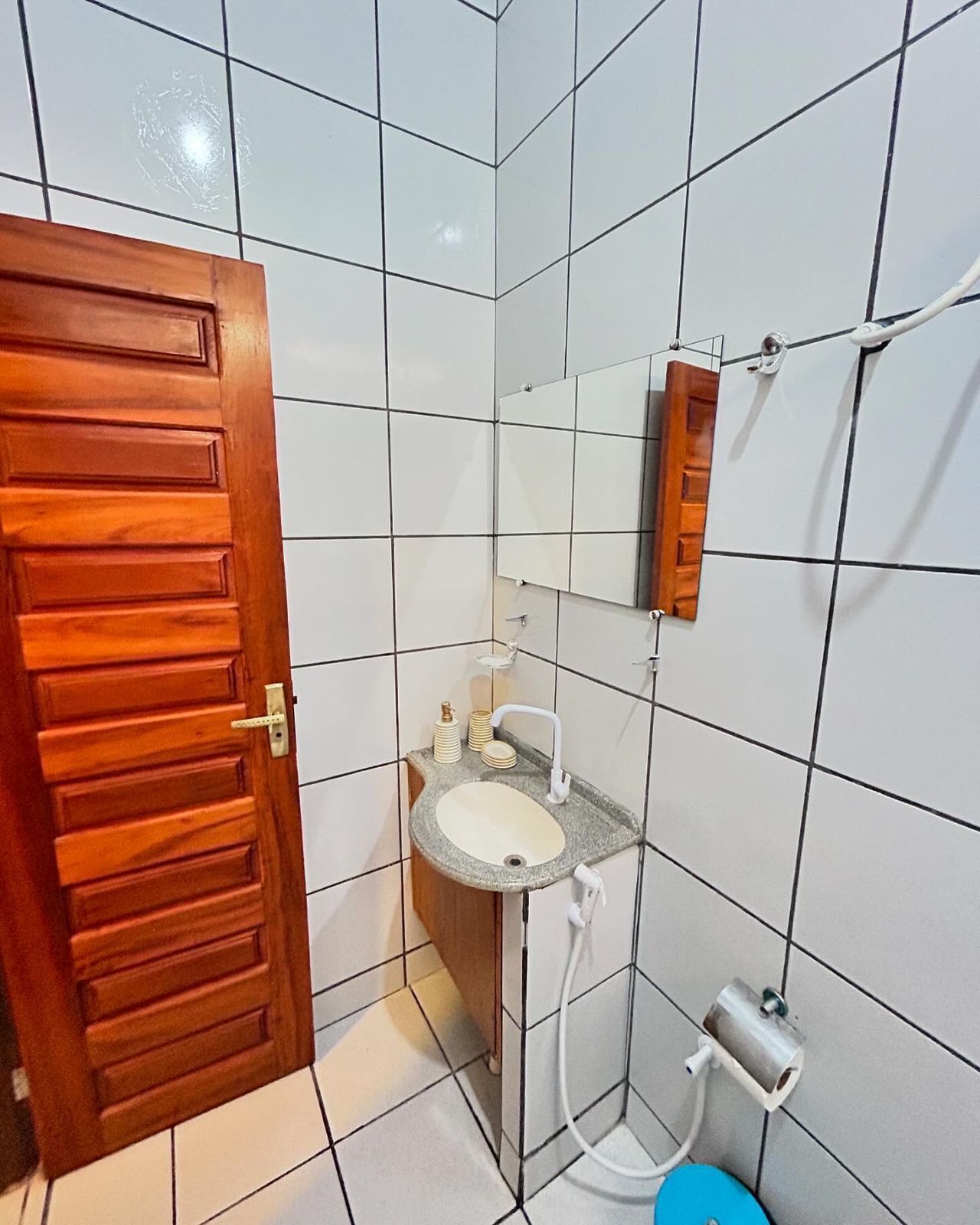 Een badkamer met witte betegelde wanden, een houten deur, een wastafel met een spiegel erboven en een toilet met een blauwe zitting.