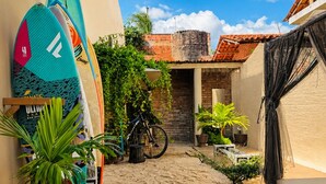 A narrow sandy pathway leading to a building with a tiled roof, surrounded by potted plants and surfboards. - Beach House Suítes Jeri (Jijoca de Jericoacoara)