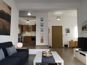 Ein modernes Wohnzimmer mit einer Couch, einem Couchtisch und einem Fernseher. Im Hintergrund ist eine Küche mit Schränken und einer Mikrowelle zu sehen.