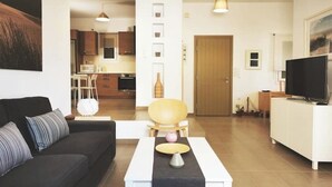 Un salon moderne avec un canapé, une table basse et un téléviseur. Au fond, on aperçoit une cuisine équipée de placards et d’un four à micro-ondes.