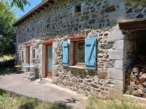 Un edificio de piedra con puerta de madera y contraventanas azules, una pequeña mesa y sillas en el exterior.