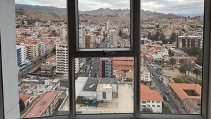 Une vue sur la ville, encadrée par une fenêtre, avec des bâtiments, des routes et des montagnes lointaines en arrière-plan.