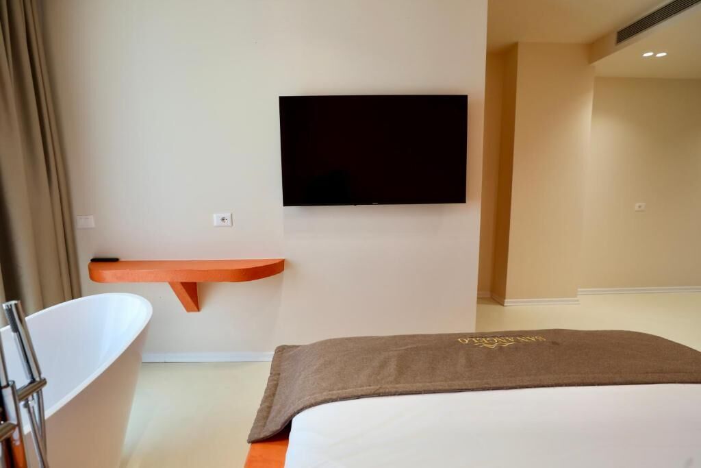 A modern hotel room with a flat-screen TV mounted on the wall, a white bathtub, and a bed with a brown blanket.
