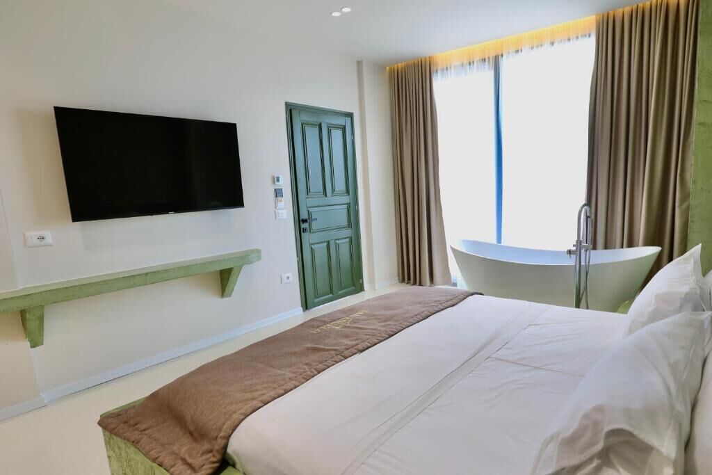 Een hotelkamer met een bed, een aan de muur bevestigde televisie, een groene deur, een raam met gordijnen en een kleine tafel.