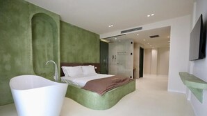 Une chambre à coucher moderne avec une grande baignoire blanche, un lit avec du linge de lit blanc et un mur d’accent vert.