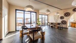 Une pièce spacieuse avec une grande table à manger, de nombreuses chaises, des luminaires suspendus et une vue dégagée sur l’extérieur grâce à de grandes fenêtres.
