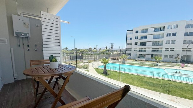 Un balcón con una mesa y sillas de madera, una pequeña planta en maceta y vistas a una piscina y edificios de apartamentos.