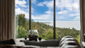 Una habitación con un ventanal grande que da a un paisaje pintoresco, una mesa con un vaso que contiene objetos, y una cama con cabecera.