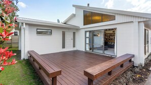 Ein modernes Haus mit einer Holzterrasse, einem großen Fenster und einer Tür, die zu einem Innenraum mit einer Couch und Bücherregalen führt.
