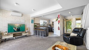 Ein modernes Wohnzimmer mit Fernseher, einem schwarzen Ledersessel, einer Küche im Hintergrund und einem Balkon mit Aussicht.