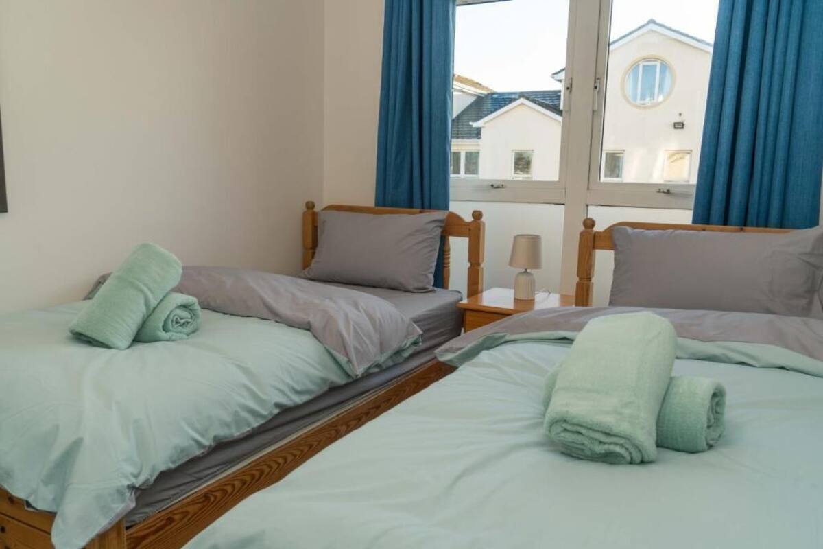 A bedroom with two beds, a window with curtains, and a bedside table with a lamp.