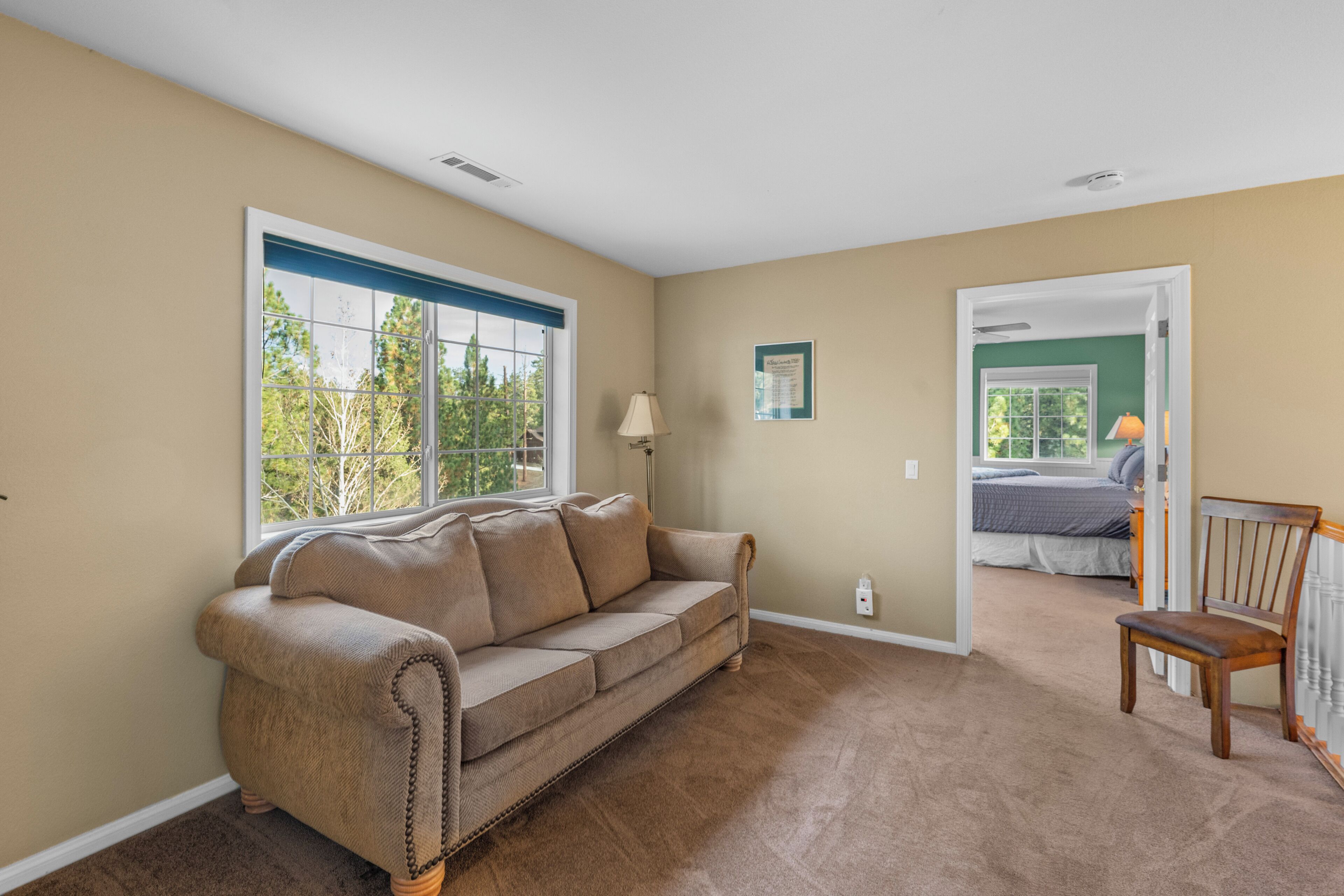 Una sala de estar con un sofá color beige, una silla de madera, una lámpara y una ventana con vista a árboles.