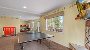 Une pièce avec une table de ping-pong, des lits superposés, une grande fenêtre donnant sur la forêt et un mur en pierre.