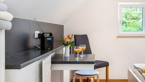 A compact kitchen with a radio, a small table with two glasses of orange juice, a chair, and a window with a view of greenery. - Guest House 'Kleines Zimmer Am See' with Shared Garden and Wi-Fi (Bodman-Ludwigshafen)