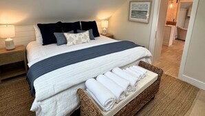 A bedroom with a skylight, a bed with a blue and white striped bedspread, two bedside lamps, a wicker basket with towels, and a view of a bathroom through an open door. - Ocean light Deck + Patio • Central P-Town (Provincetown)