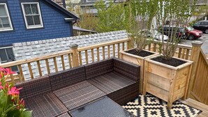 A deck with a brown wicker sofa, a patterned rug, and potted plants. - Ocean light Deck + Patio • Central P-Town (Provincetown)