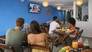 A dining area with wooden tables and chairs, a television mounted on the wall, and a blue wall. - Pousada Pé no Mar (Japaratinga)