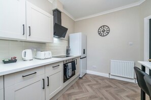 A compact kitchen with white cabinets, a built-in oven, a white refrigerator, a clock, and a radiator.