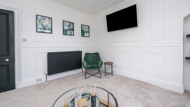 A room with a black door, white walls, a green armchair, a small round table, and a mounted flat-screen TV. - The Blue Toon Retreat | 3 Bed Apartment (Aberdeenshire)