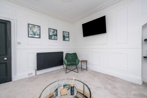A room with a black door, white walls, a green armchair, a small round table, and a mounted flat-screen TV. - The Blue Toon Retreat | 3 Bed Apartment (Aberdeenshire)