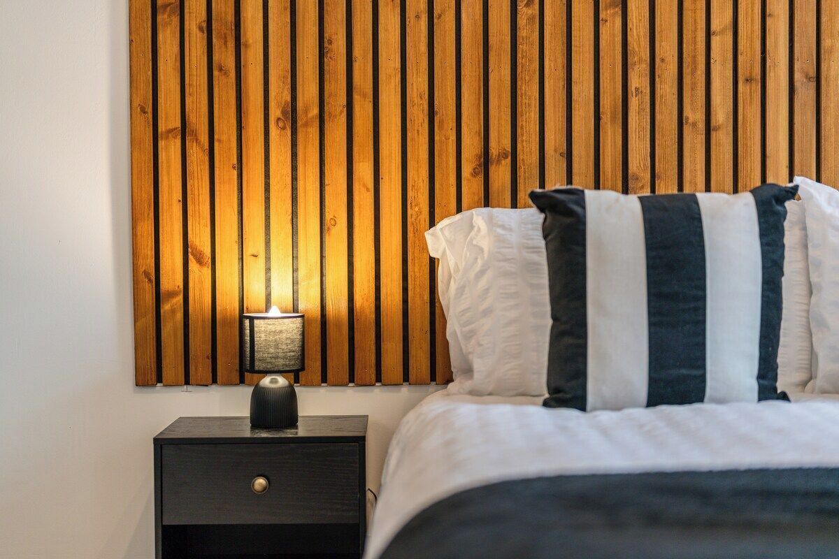 Un dormitorio con cabecera de madera con listones, una mesita de noche con lámpara, y una cama con almohadas a rayas.