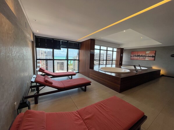 A modern bathroom with a large bathtub, two red lounge chairs, and a view of the city through large windows. - Apartamento no Edifício Time Ap 1105 (Maceió)