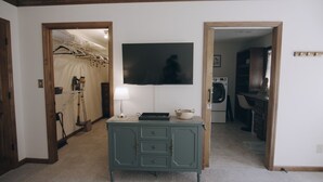 Una habitación con escritorio, televisión de pantalla plana, lavadora y un pasillo que conduce a un armario con ropa colgada.