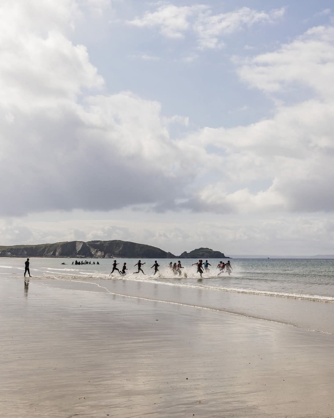 A beach with people running along the shore, a person walking, and distant hills.