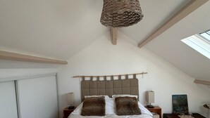 Een slaapkamer met een hoog plafond, een geweven lamp, een bed met witte lakens en bruine kussens, een nachtkastje met een lamp, en een schilderij aan de muur.