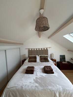 Ein Schlafzimmer mit hohen Decken, einer gewobenen Leuchte, einem Bett mit weißen Leinen und braunen Kissen, einem Nachttisch mit Lampe und einem Bild an der Wand.