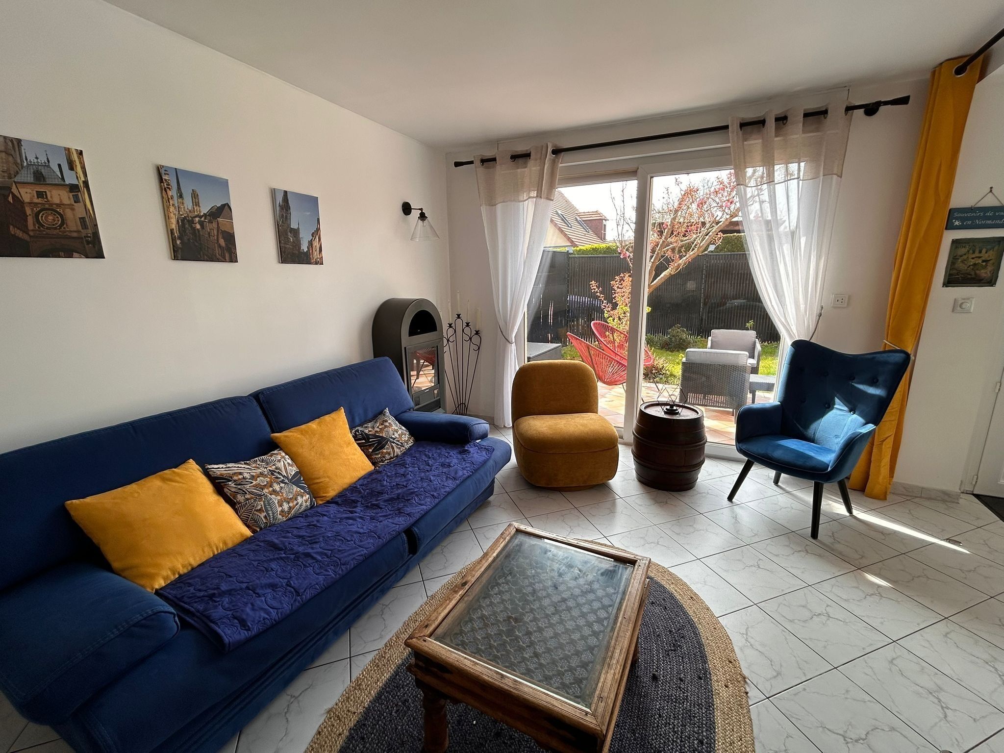 A living room with a blue sofa, a yellow armchair, a glass coffee table, and a view of the outside.