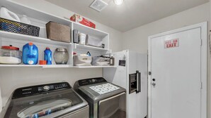 Ein Waschraum mit einer Samsung Waschmaschine und einem Trockner, Regalen mit Reinigungsmitteln und einem Kühlschrank.