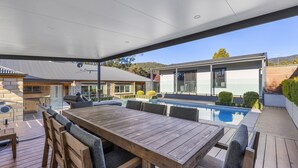 A modern outdoor patio with a wooden table and chairs, a swimming pool, and a contemporary house in the background. - Route 86 Bright stunning holiday home with private pool. (Bright)
